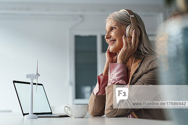 Smiling senior businesswoman listening music on headphones sitting at desk in office