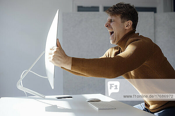 Angry businessman lifting computer monitor screaming at desk in office
