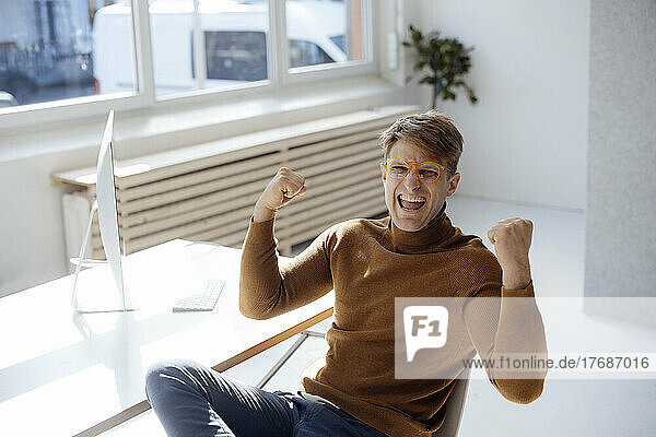 Happy businessman wearing eyeglasses gesturing fist sitting at desk in office