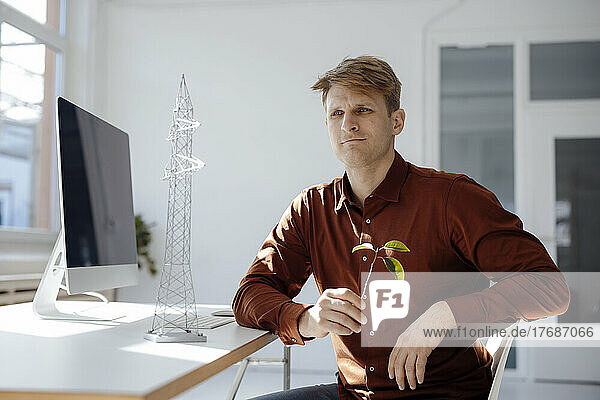 Thoughtful engineer holding leaf sitting by electricity pylon model at desk in office