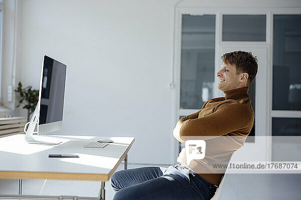 Smiling businessman sitting with arms crossed looking at computer in office