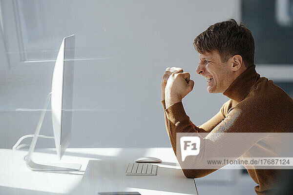 Happy businessman gesturing fist looking at desktop PC in office