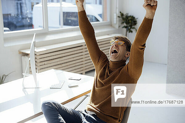 Happy businessman with arms raised shouting sitting at desk in office