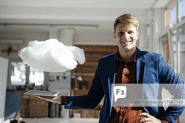 Happy businessman holding tablet PC under levitating cloud network in office