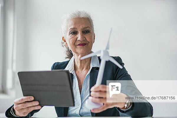 Lächelnde Geschäftsfrau mit Windturbinenmodell und Tablet-PC sitzt am Schreibtisch im Büro
