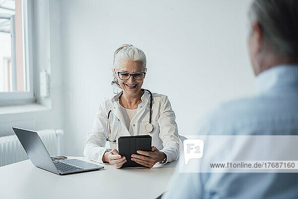 Happy doctor using tablet PC sitting with patient at desk