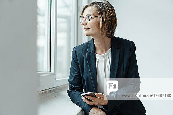 Lächelnde Geschäftsfrau mit Smartphone  die aus dem Fenster schaut