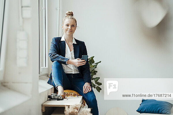 Businesswoman with smart phone sitting near window at office