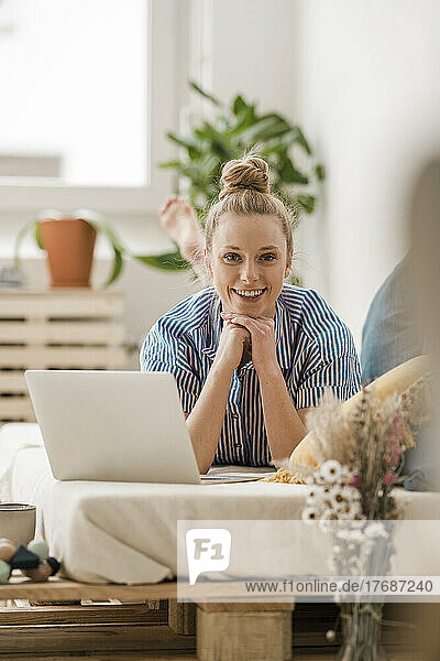 Smiling businesswoman with hand on chin lying by laptop on bed at home