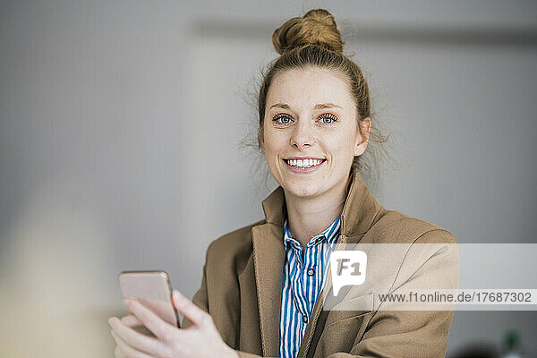 Smiling businesswoman wearing blazer holding smart phone at office