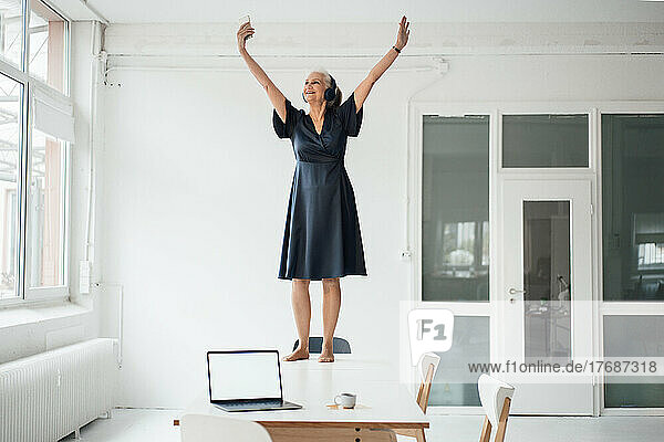 Happy businesswoman with arms raised standing on desk in office