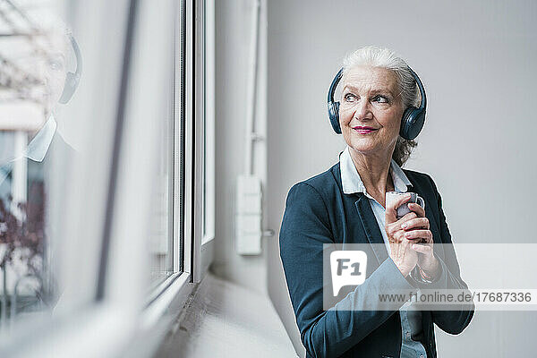 Businesswoman wearing wireless headphones standing by window in office