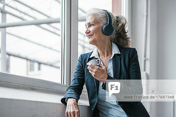 Smiling businesswoman listening music through wireless headphones standing by window in office