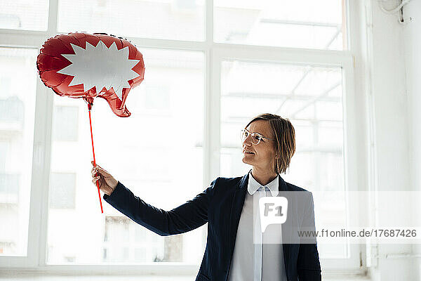 Smiling businesswoman with speech bubble balloon in front of window at office