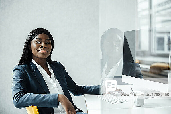 Smiling young businesswoman sitting with desktop PC at desk seen through glass