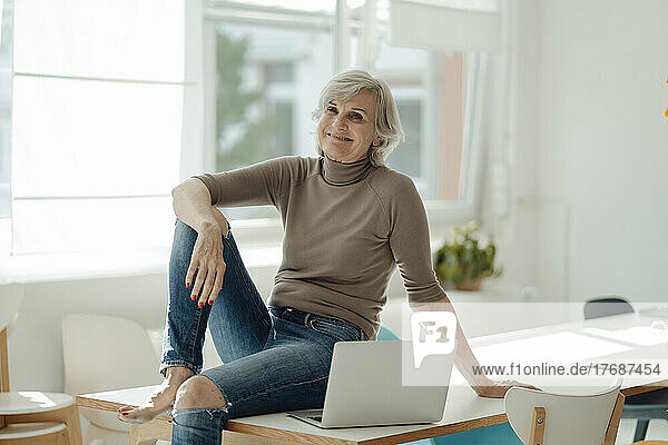 Smiling businesswoman with laptop sitting on desk in office