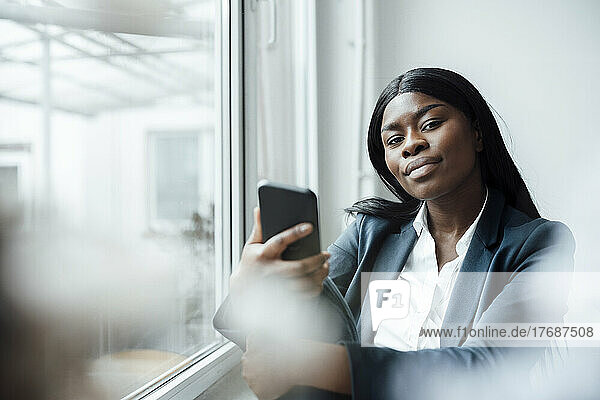 Businesswoman holding smart phone sitting by window in office