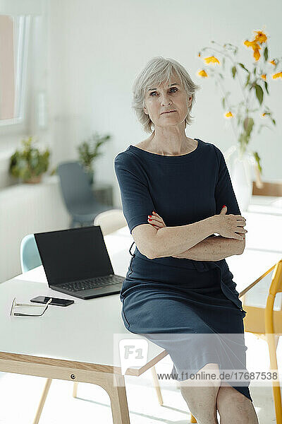 Senior businesswoman with arms crossed sitting on desk in office
