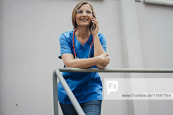 Smiling doctor talking through smart phone leaning on railing in front of wall