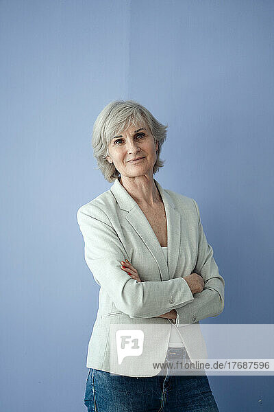 Smiling senior businesswoman with arms crossed standing against blue background