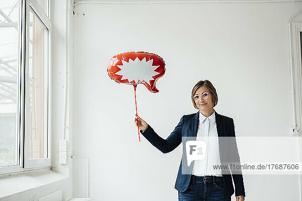 Smiling businesswoman with speech bubble balloon in front of wall at office
