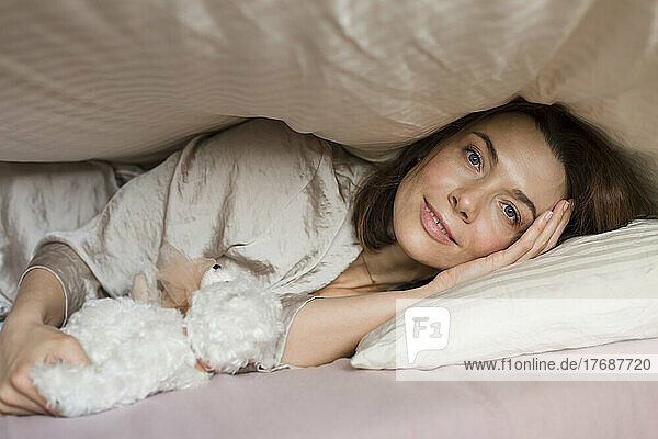 Smiling mature woman with teddy bear toy lying under blanket on bed at home