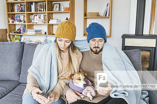 Couple with dog sitting on couch wrapped in blanket looking at smartphone