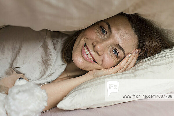Happy mature woman with teddy bear toy lying under blanket on bed at home