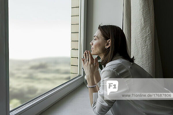 Mature woman with hands clasped leaning on window sill at home