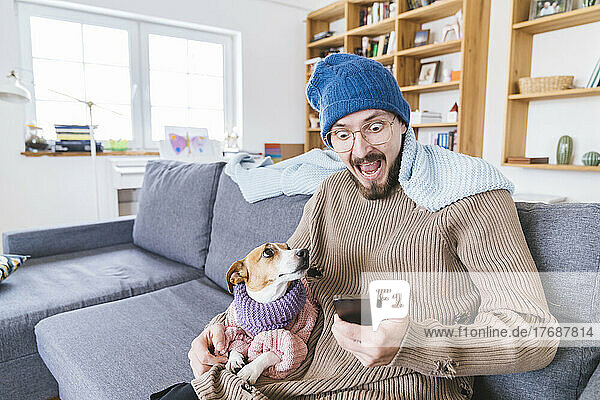 Schockierter Mann mit Wollmütze sitzt auf Couch und hält Hund in der Hand und checkt Smartphone