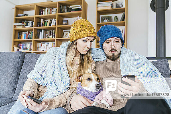 Paar mit Hund sitzt in Decke gehüllt auf Couch und schaut aufs Smartphone