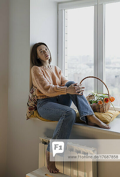 Mature woman sitting on window sill with basket of tulips at home