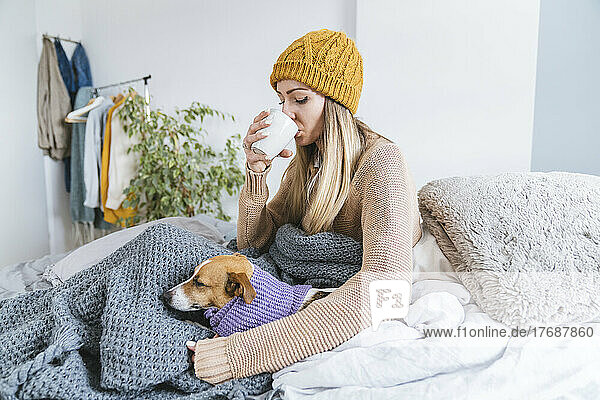 Young woman wearing wooly hat drinkig hot tea sitting in bed with dog
