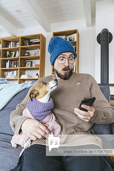 Schockierter Mann mit Wollmütze sitzt auf Couch und hält Hund in der Hand und checkt Smartphone