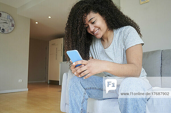 Smiling young woman using smart phone sitting on sofa at home