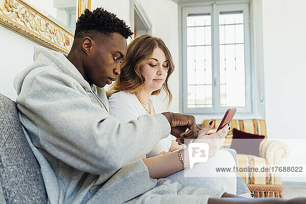 Gemischtrassiges Paar nutzt gemeinsam sein Smartphone und sitzt zu Hause auf dem Sofa