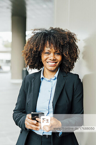 Happy Afro businesswoman holding mobile phone leaning on wall