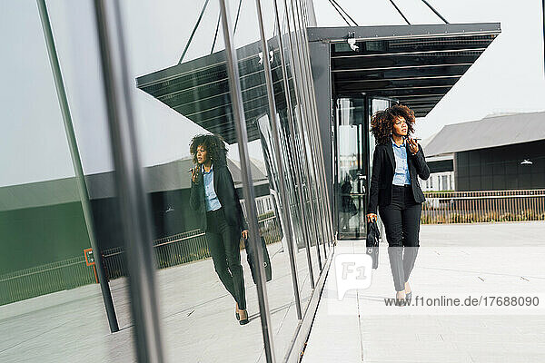 Businesswoman holding laptop bag talking on mobile phone through speaker walking by reflection