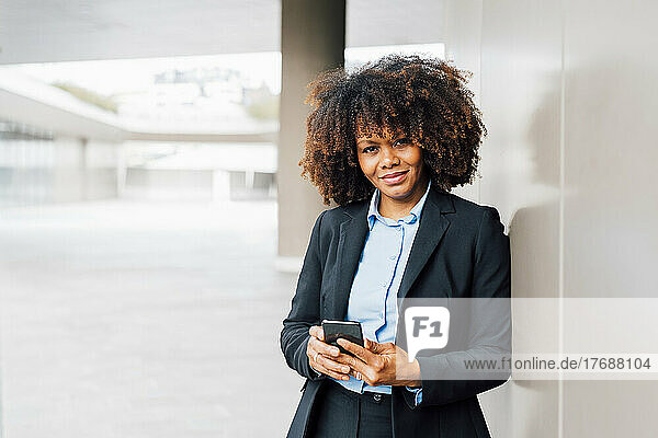 Lächelnde Afro-Geschäftsfrau hält ihr Mobiltelefon an die Wand gelehnt