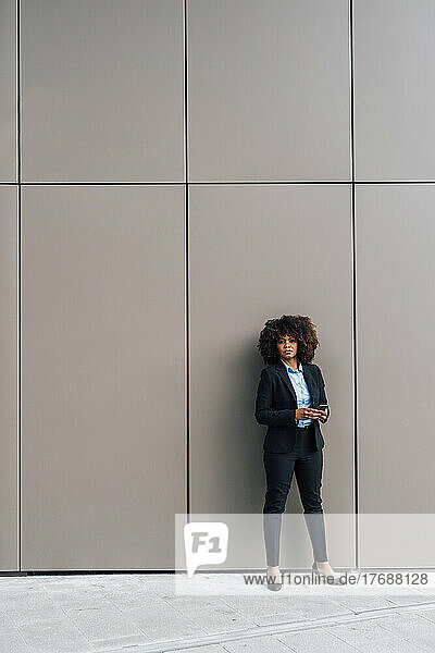 Businesswoman holding mobile phone standing in front of wall