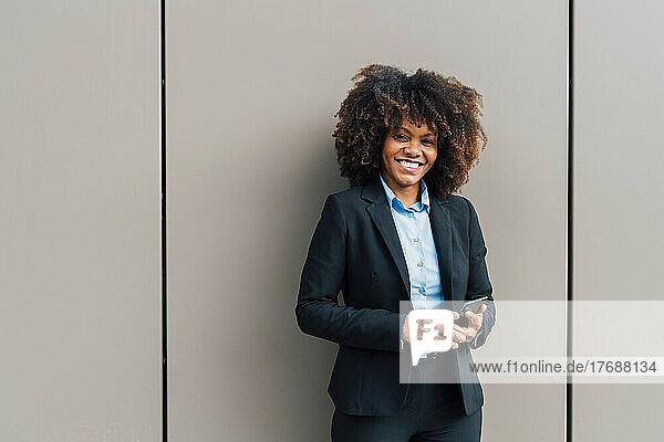 Happy Afro businesswoman holding mobile phone standing in front of wall