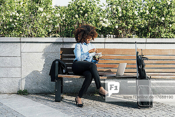 Smiling businesswoman eating salad sitting with laptop on bench