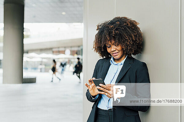 Smiling Afro businesswoman using smart phone leaning on wall