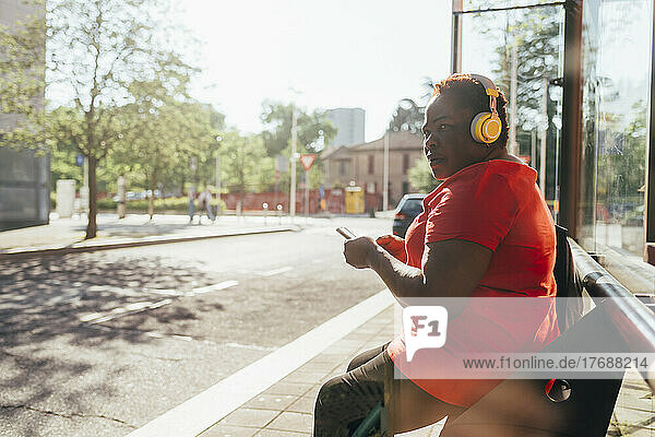 Woman holding smart phone listening music through wireless headphones sitting on bench