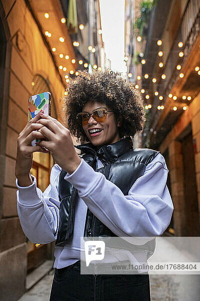 Glückliche junge Frau mit Sonnenbrille  die in einer Gasse ein Selfie mit dem Smartphone macht