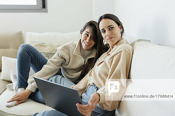 Smiling young woman sitting with laptop by girlfriend on sofa in living room at home