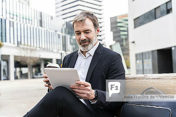 Businessman holding disposable coffee cup using tablet PC sitting on bench