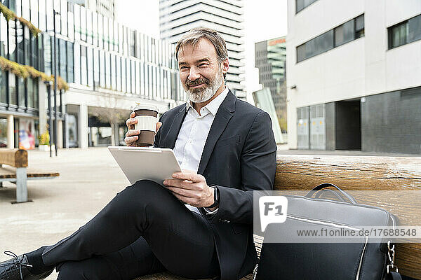 Smiling mature businessman with tablet PC and disposable coffee cup sitting on bench