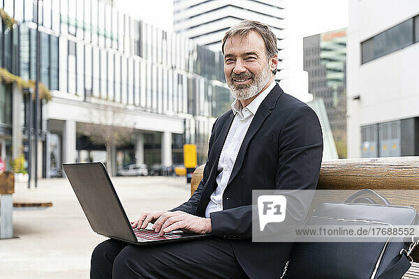 Smiling businessman with laptop sitting on bench