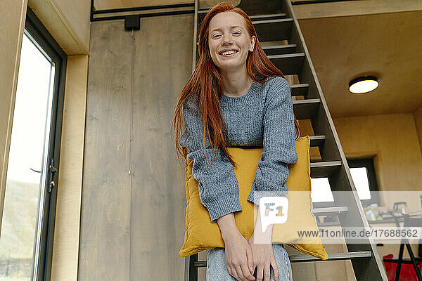 Lächelnde junge Frau mit Kissen sitzt auf Leiter